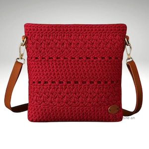 以下が含まれることがあります： 茶色の革のストラップと金色の金具が付いた、赤いかぎ針編みのクロスボディバッグ。バッグはテクスチャードステッチパターンと小さな楕円形の革タグが特徴です。ストラップは調節可能で、バッグは長方形です。