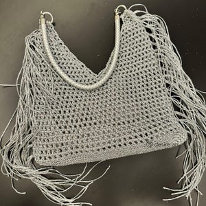 May include: A gray crocheted handbag with fringe detailing. The bag features a silver braided handle and silver fringe along the edges. The open weave design and fringe give the bag a bohemian aesthetic.