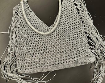 May include: A gray crocheted handbag with fringe detailing. The bag features a silver braided handle and silver fringe along the edges. The open weave design and fringe give the bag a bohemian aesthetic.