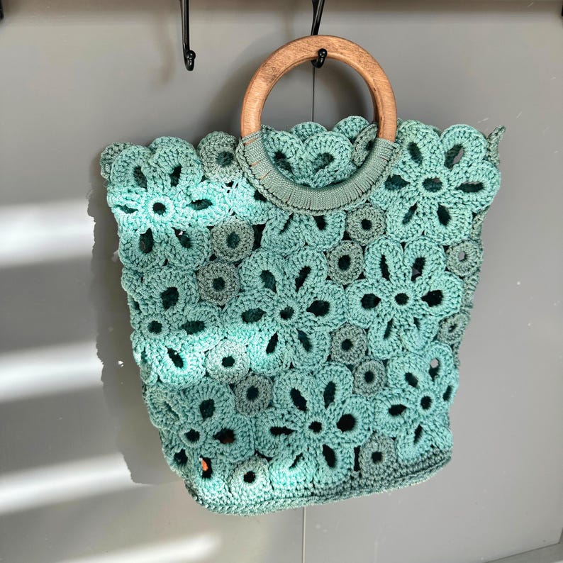 May include: A light green crocheted tote bag with a wooden handle. The bag is made of a delicate, floral pattern.