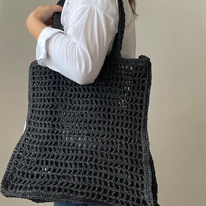 Könnte beinhalten: Eine schwarze gehäkelte Tragetasche mit dicken Henkeln. Die Tasche ist aus gewebtem Material und hat eine große, offene Oberseite.