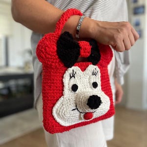 Puede incluir: Bolso de crochet rojo con una cara blanca y orejas y lazo negros. La cara tiene ojos negros, una nariz negra y una boca roja.