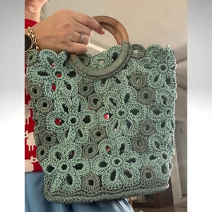 May include: A crocheted tote bag with a wooden handle. The bag is made of a light green yarn and features a floral pattern.