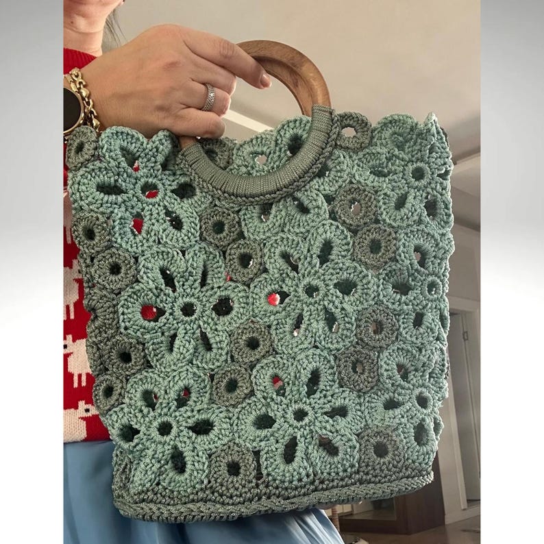 May include: A crocheted tote bag with a wooden handle. The bag is made of a light green yarn and features a floral pattern.