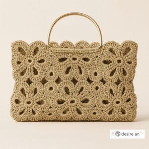 Op de afbeelding: Een beige gehaakte handtas met een goudkleurige handgreep. De tas heeft een bloemenpatroon over de rechthoekige body en een geschulpte rand. De handgreep is een eenvoudige, afgeronde staaf.
