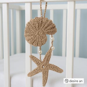 Op de afbeelding: Een handgemaakte, beige gehaakte mobiel met een schelp- en zeesterontwerp, versierd met drie imitatieparels. De mobiel is opgehangen aan een gevlochten koord en is ontworpen om aan een wieg of een vergelijkbare structuur te hangen. Het thema is nautisch en kust.