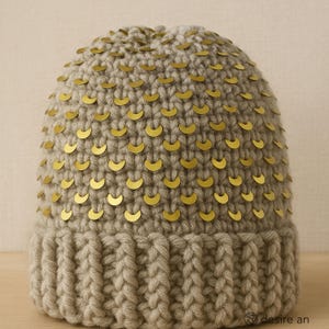 May include: A knitted beanie in a neutral beige color, adorned with numerous gold-colored crescent-shaped sequins. The hat features a ribbed brim and a textured, handcrafted appearance, suitable for cold weather.