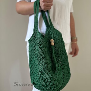 Könnte beinhalten: Eine grüne gehäkelte Tragetasche mit einem Holzperlen-Quaste. Die Tasche hat ein geometrisches Muster und wird von einer Person gehalten.