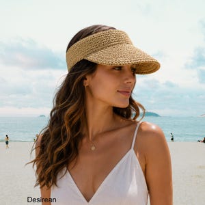 May include: A beige woven straw visor with a wide brim and a textured, crocheted appearance. The visor features a structured front panel and a band that wraps around the head. The word Desirean is printed in the bottom left corner of the image.