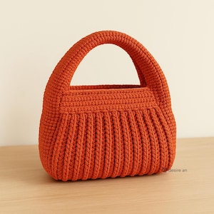以下が含まれることがあります： 丸いハンドルが付いたオレンジ色のかぎ針編みのハンドバッグ。バッグの下部にはリブ編みのテクスチャがあり、上部は滑らかです。バッグは明るい茶色の表面に置かれています。