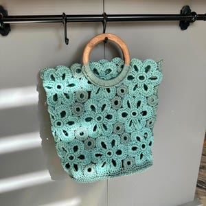 May include: A teal crocheted bag with a wooden handle. The bag is made of a delicate floral pattern.