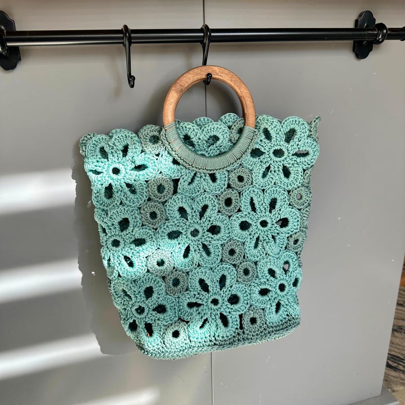May include: A teal crocheted bag with a wooden handle. The bag is made of a delicate floral pattern.