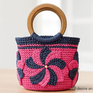 May include: A pink and navy blue crocheted handbag with a wooden handle. The bag features a floral design with pearl accents. The handle is made of light brown wood. The bag is a stylish accessory.