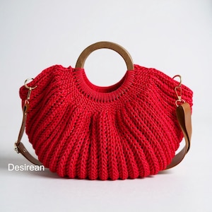 Puede incluir: Un bolso de mano rojo vibrante con un asa de madera y una correa de cuero marrón desmontable. El bolso está hecho de un material tejido texturizado y presenta un asa de madera circular. La palabra "Desirean" está impresa en la parte inferior izquierda.