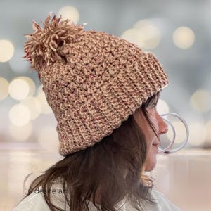 以下が含まれることがあります： ピンクとブラウンの色の組み合わせの、ふわふわのポンポンが付いた、かぎ針編みニットの帽子