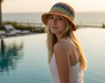 May include: A multi-colored crochet bucket hat with a wide brim. The hat features a pattern of orange, green, and blue stripes. The hat is worn by a person with long blonde hair, and is set against a blurred background of water and sky.