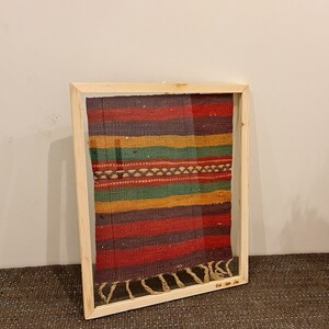 May include: A framed piece of colorful woven fabric with a fringe on the bottom. The fabric is a mix of red, green, yellow, and brown stripes.