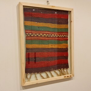 May include: A framed tapestry with a colorful striped pattern in shades of red, brown, green, and yellow. The tapestry has a fringe at the bottom.