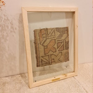 May include: A framed wall decor rug with a geometric pattern in shades of brown and beige. The frame is made of natural wood and has a glass front. The text "WALL DECOR RUG" is printed on a gold label at the bottom of the frame.