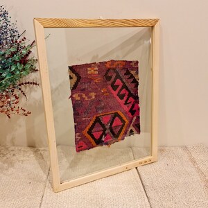 May include: A framed piece of colorful fabric with a geometric pattern. The fabric is a mix of pink, purple, orange, and black. The frame is made of light wood and has a clear glass front.