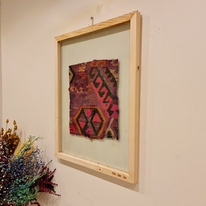 May include: A framed piece of colorful woven fabric with a geometric pattern. The frame is made of natural wood and has a glass front. The fabric is a mix of red, pink, black, and white.