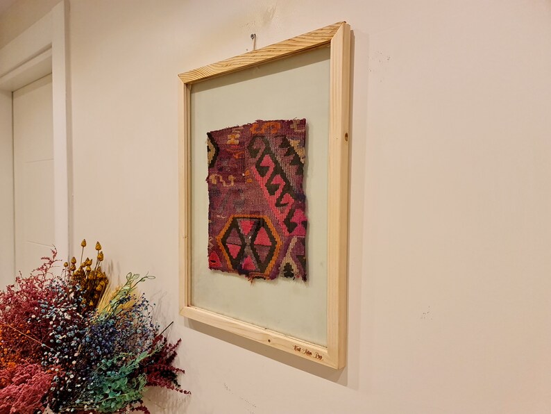 May include: A framed piece of colorful woven fabric with a geometric pattern. The frame is made of natural wood and has a glass front. The fabric is a mix of red, pink, black, and white.