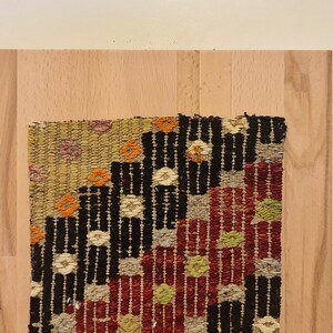Puede incluir: Un tapiz tejido colorido con patrones geom&eacute;tricos en tonos rojos, negros, blancos y verdes, enmarcado en un marco de madera claro.
