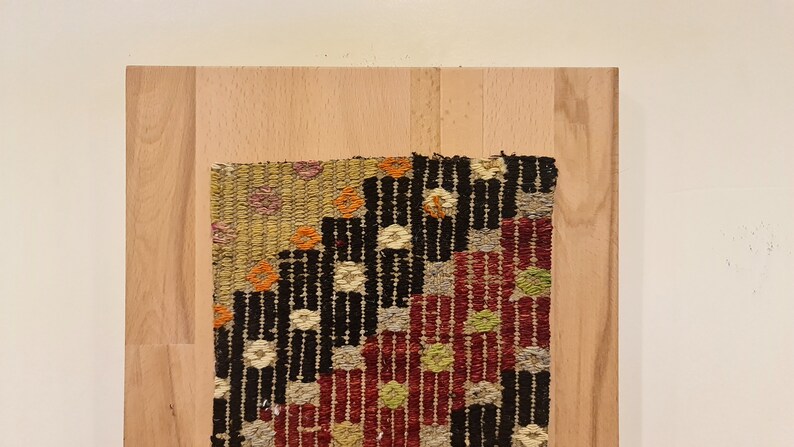 Puede incluir: Un tapiz tejido colorido con patrones geom&eacute;tricos en tonos rojos, negros, blancos y verdes, enmarcado en un marco de madera claro.