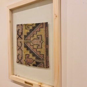 May include: A framed wall hanging with a colorful woven rug inside. The rug has a geometric pattern in shades of pink, yellow, black, and brown. The frame is made of light wood and has a brass plaque that reads "WALL OULM RUG".