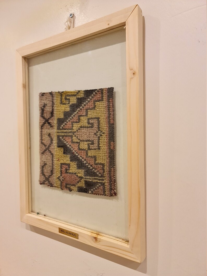 May include: A framed wall hanging with a colorful woven rug inside. The rug has a geometric pattern in shades of pink, yellow, black, and brown. The frame is made of light wood and has a brass plaque that reads "WALL OULM RUG".