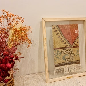 May include: A framed vintage rug with a geometric pattern in shades of red, yellow, blue, and green. The rug has fringe on the bottom and is displayed in a wooden frame with a glass front.