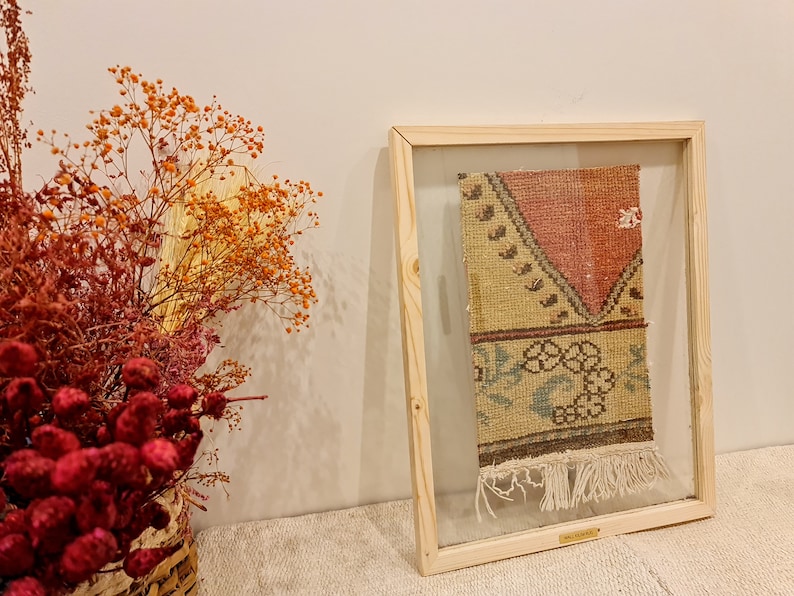 May include: A framed vintage rug with a geometric pattern in shades of red, yellow, blue, and green. The rug has fringe on the bottom and is displayed in a wooden frame with a glass front.