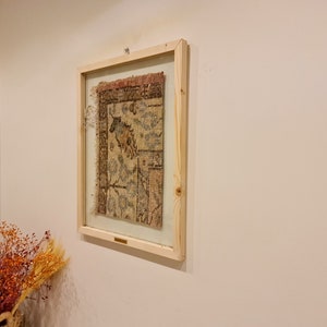 May include: A framed piece of antique fabric with a faded floral pattern in shades of brown, blue, and white. The fabric is displayed behind glass in a simple wooden frame.