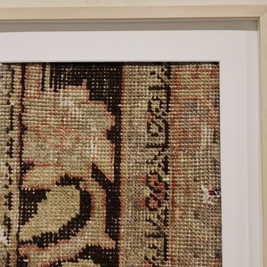 K&ouml;nnte beinhalten: Ein gerahmter Vintage-Teppich mit einem detaillierten Blumenmuster in Braun-, Beige- und Rosat&ouml;nen. Der Teppich ist in einem einfachen Holzrahmen mit einer wei&szlig;en Passepartout gerahmt.