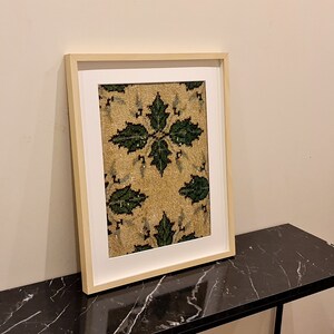 May include: A framed artwork with a beige background and a green floral pattern. The frame is a light wood colour and the artwork is hanging on a white wall.