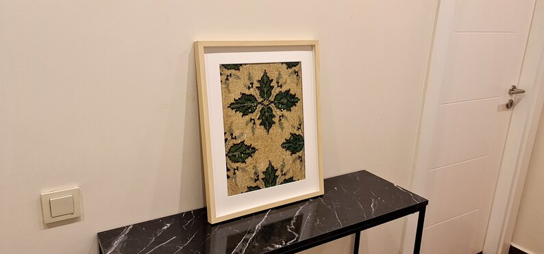 May include: A framed artwork with a beige background and a green floral pattern. The frame is a light wood colour and the artwork is hanging on a white wall.