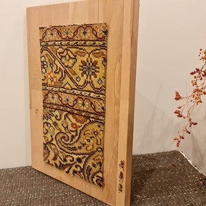 May include: A rectangular wooden frame with a colorful patterned fabric mounted inside. The fabric has a floral design with shades of yellow, brown, and red. The frame is standing on a brown surface.