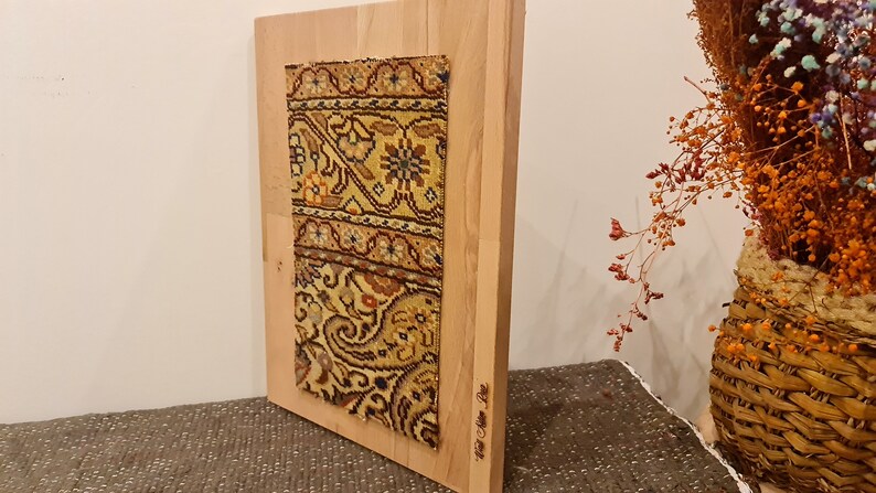 May include: A rectangular wooden frame with a colorful patterned fabric mounted inside. The fabric has a floral design with shades of yellow, brown, and red. The frame is standing on a brown surface.