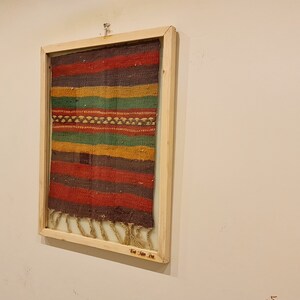 May include: A framed colorful woven tapestry with a fringe on the bottom. The frame is made of wood and has a label that reads "The Jane Doe Co."