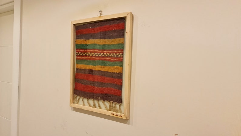 May include: A framed colorful woven tapestry with a fringe on the bottom. The frame is made of wood and has a label that reads "The Jane Doe Co."