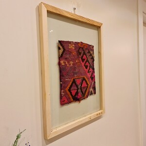 May include: A framed piece of colorful woven fabric with a geometric pattern in shades of pink, red, orange, and black. The fabric is displayed in a simple wooden frame.