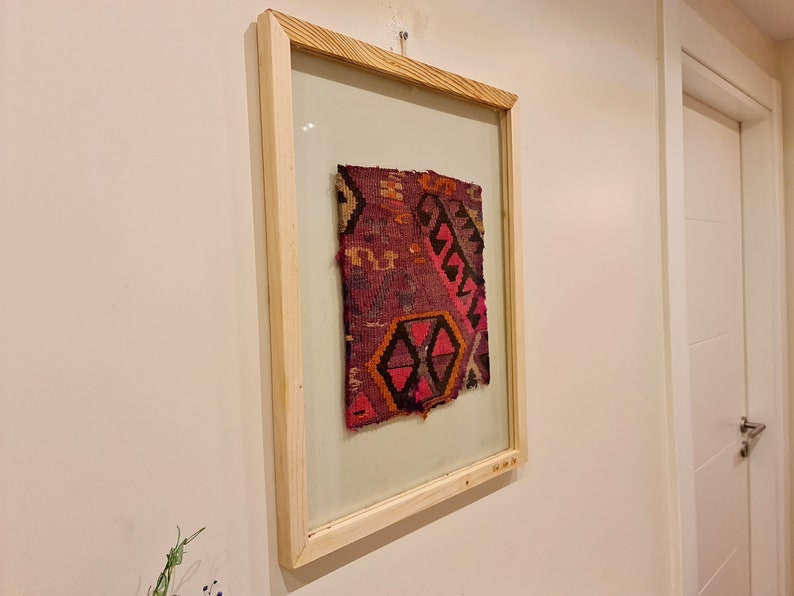 May include: A framed piece of colorful woven fabric with a geometric pattern in shades of pink, red, orange, and black. The fabric is displayed in a simple wooden frame.