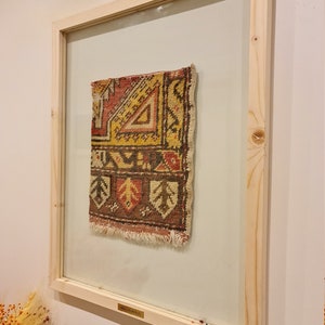 May include: A framed vintage rug with a geometric pattern in shades of red, yellow, brown, and white. The frame is made of natural wood and has a glass front. The rug is labelled "Small Rug".