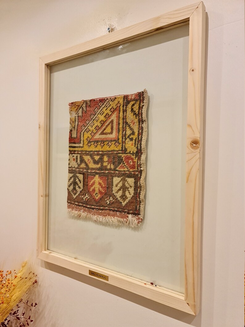 May include: A framed vintage rug with a geometric pattern in shades of red, yellow, brown, and white. The frame is made of natural wood and has a glass front. The rug is labelled "Small Rug".