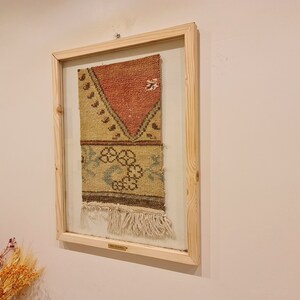 May include: A framed piece of vintage woven fabric with a geometric pattern in shades of red, brown, yellow, and blue. The fabric has a fringe border and is displayed in a wooden frame.