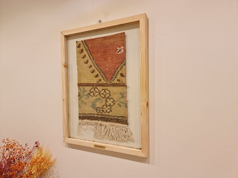 May include: A framed piece of vintage woven fabric with a geometric pattern in shades of red, brown, yellow, and blue. The fabric has a fringe border and is displayed in a wooden frame.