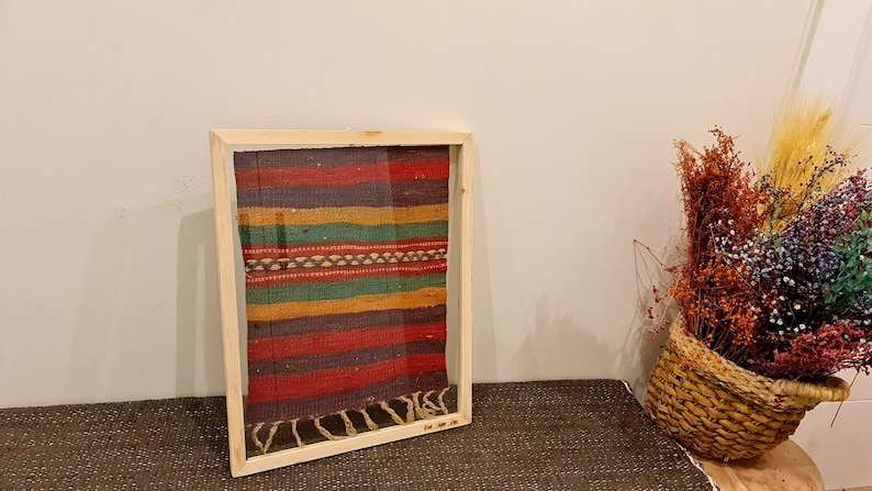 May include: A framed piece of colorful woven fabric with a fringe on the bottom. The fabric is a mix of red, green, yellow, and brown stripes.