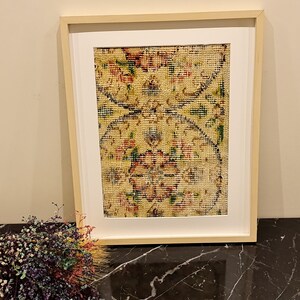 May include: A framed print of a vintage rug with a floral pattern in shades of red, yellow, green, and blue. The rug is framed in a light wood frame.