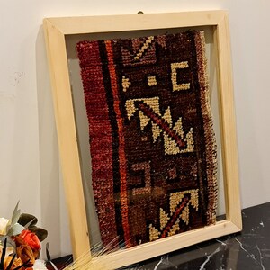 May include: A framed piece of antique fabric with a geometric pattern in shades of red, brown, and cream. The fabric is displayed in a natural wood frame with a glass front.