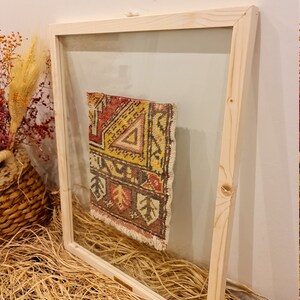 May include: A small, colourful rug with geometric patterns is framed in a natural wood frame with a clear glass front.